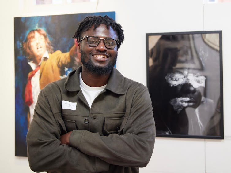 An African man smiles for the camera, arms crossed. Behind him are framed artworks on a wall.