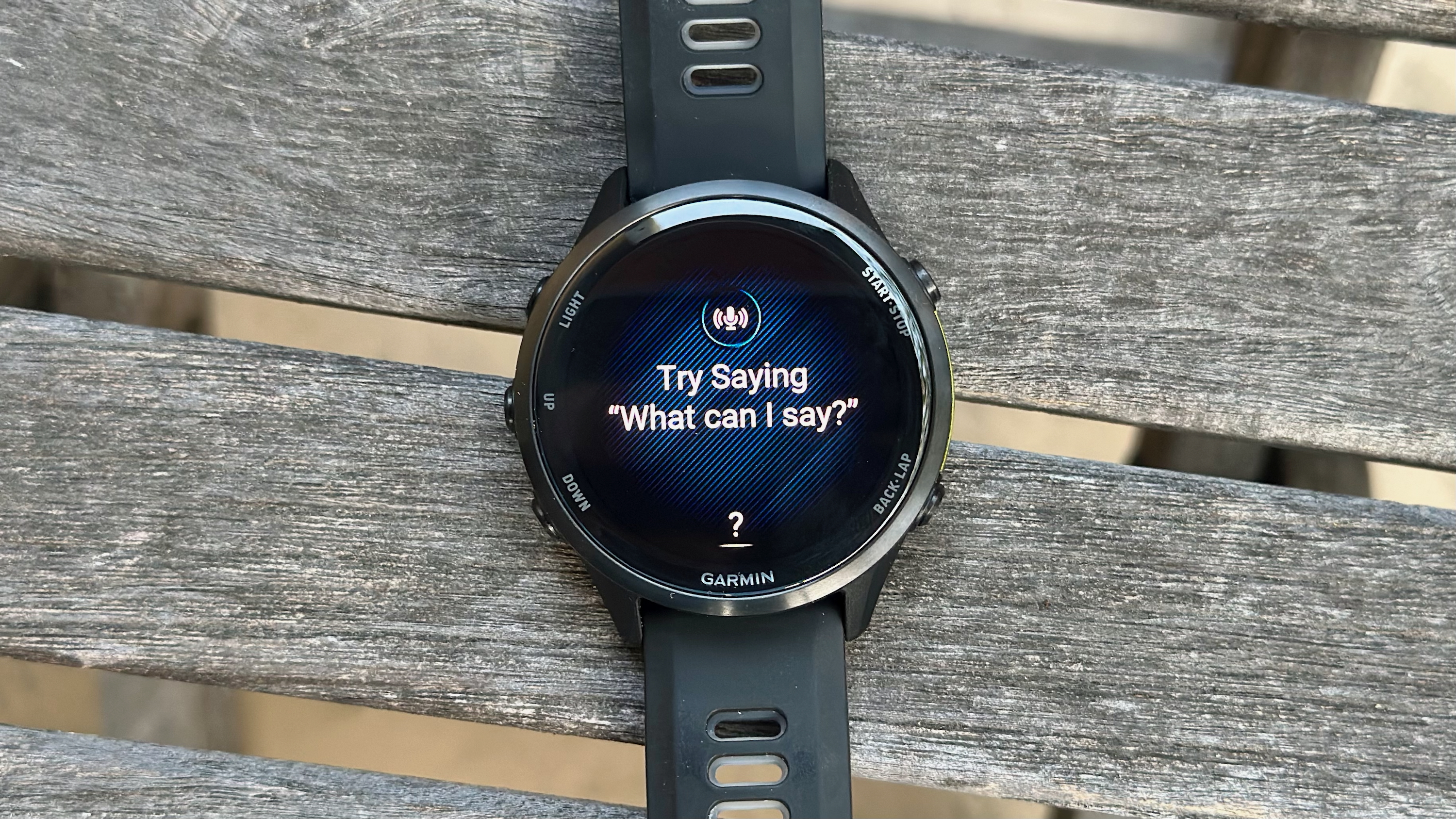The Garmin Forerunner 970 on a table outside, the display showing the text "Try Saying 'What can I say?'" for the Voice Assistant.