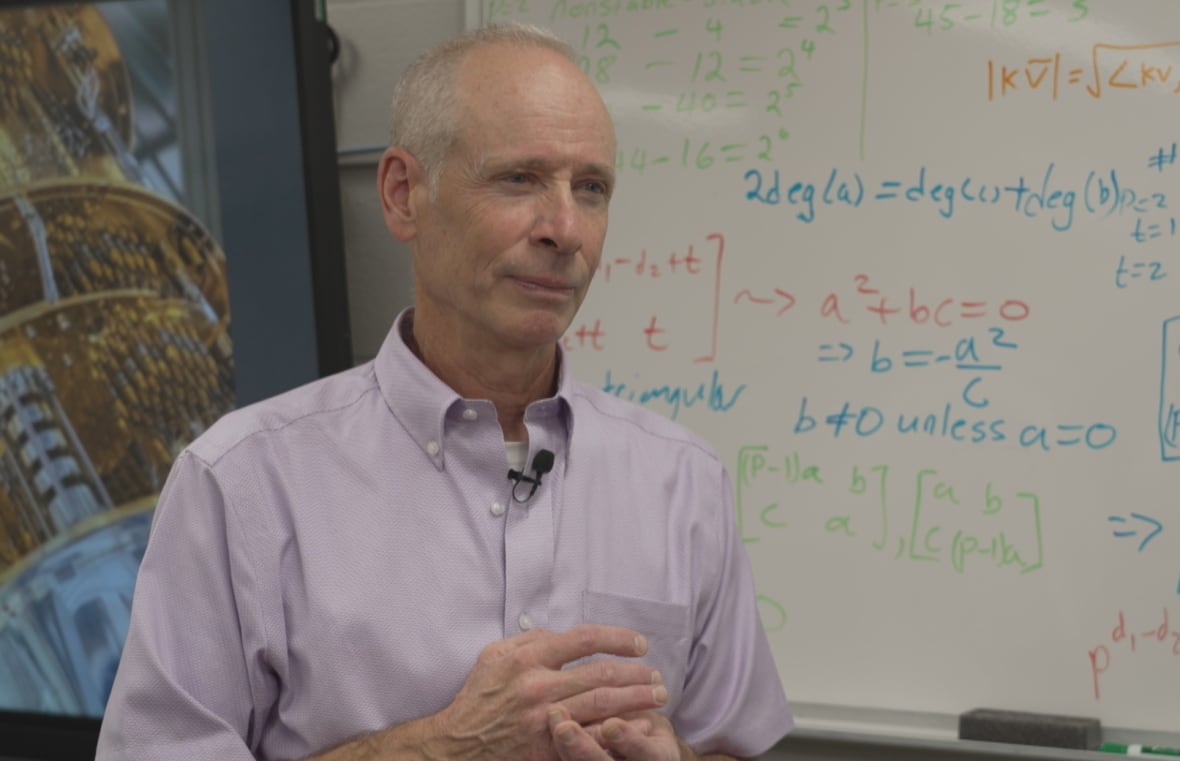 A man in a dress shirt stands in front of a white board with mathematic equations on it. 