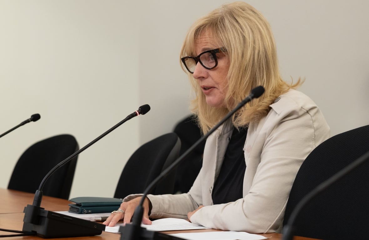 A woman with glasses and blond hair speaks into a microphone.