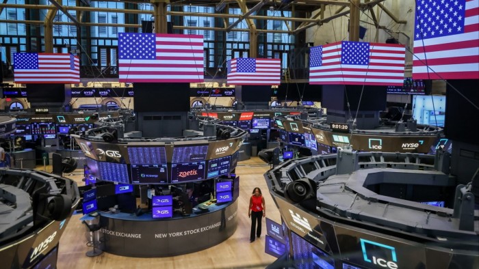 A woman walks on the trading floor of the New York Stock Exchange, surrounded by trading stations and large digital American flags.