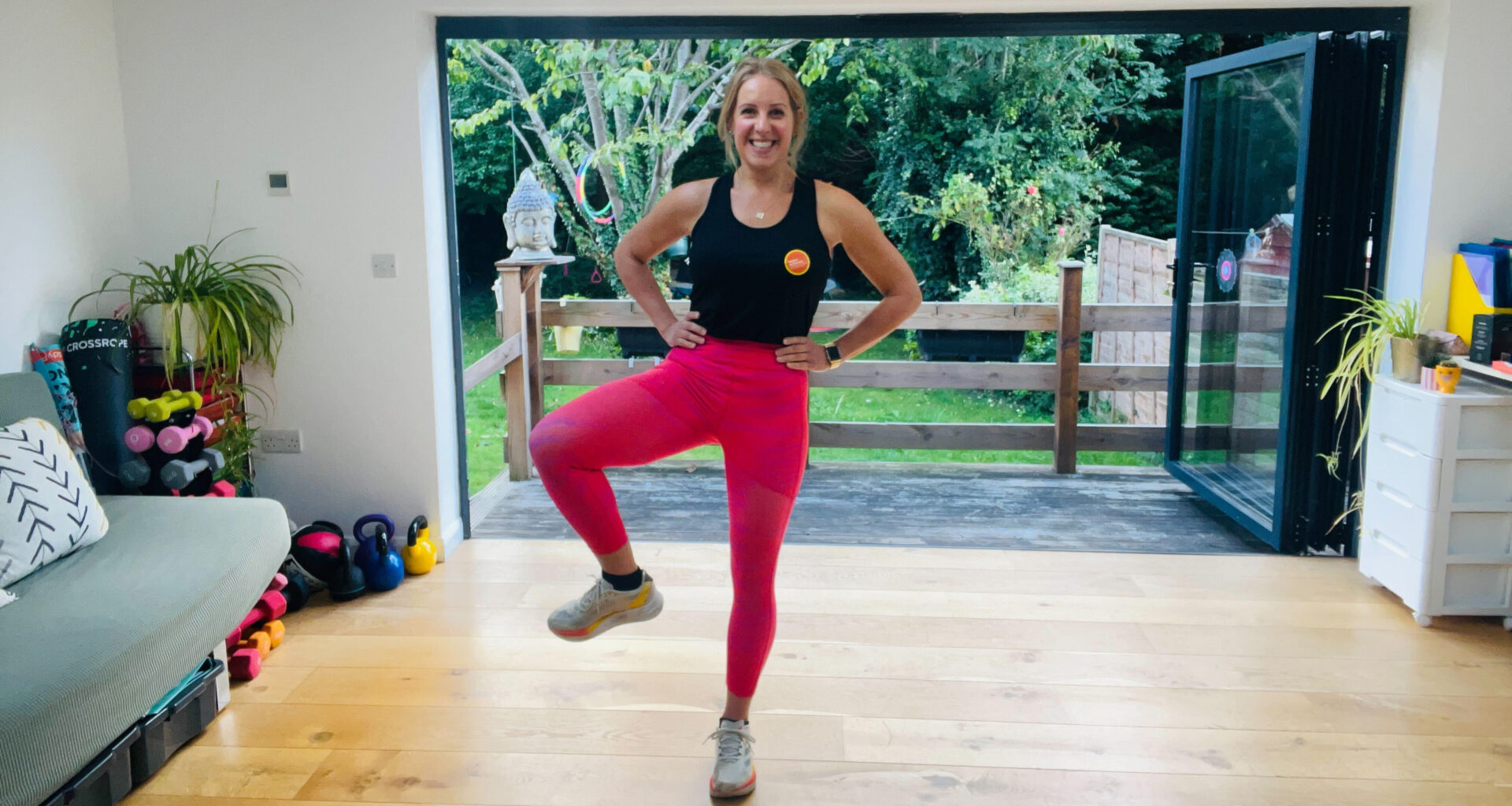 Fitness journalist Maddy Biddulph stands performing a hip opening exercise in her living room. Her hands are on her hips and her right knee is bent and held out to the side. She is smiling and behind her we see open glass doors and a green leafy garden.