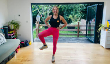 Fitness journalist Maddy Biddulph stands performing a hip opening exercise in her living room. Her hands are on her hips and her right knee is bent and held out to the side. She is smiling and behind her we see open glass doors and a green leafy garden.