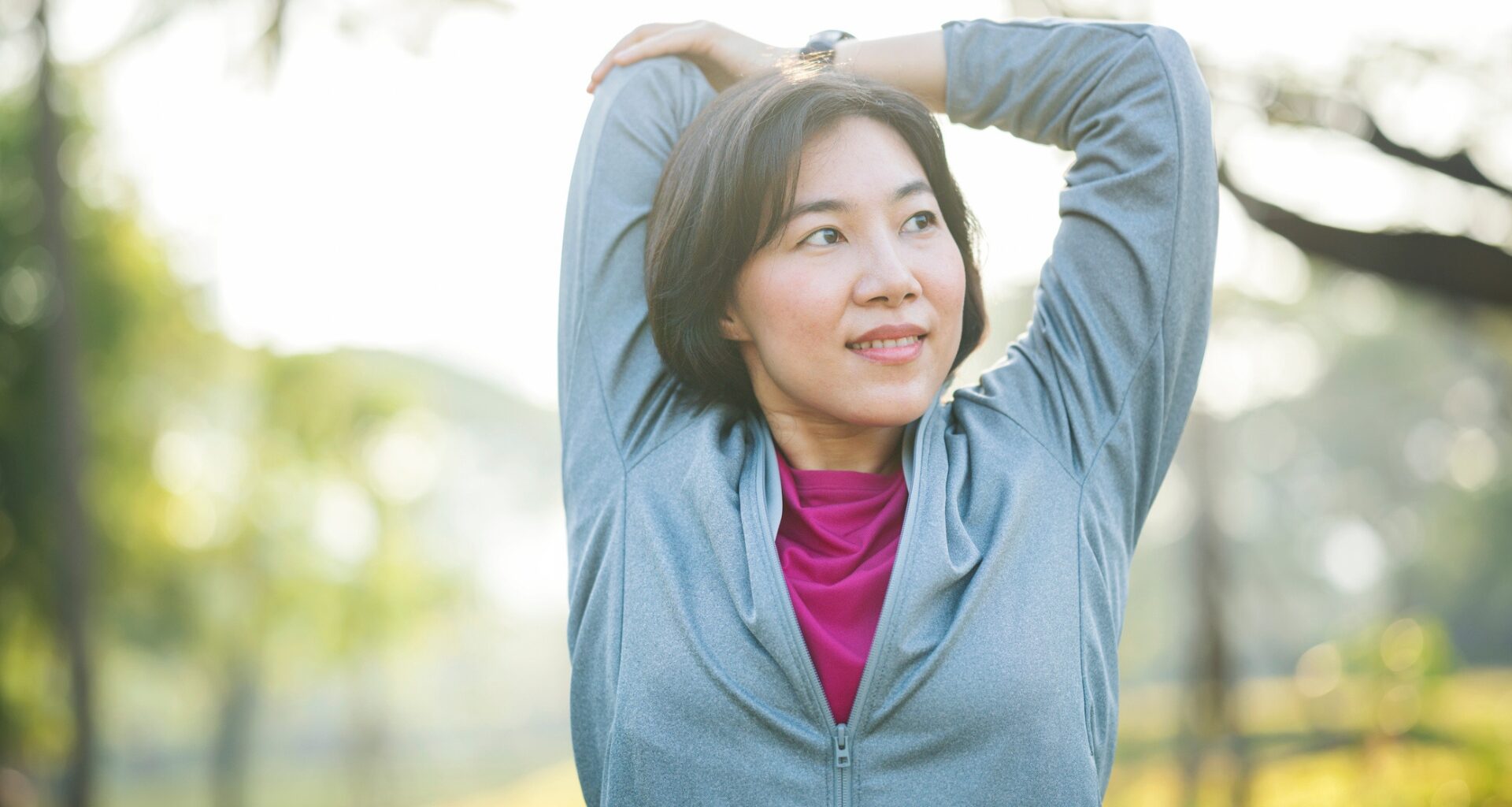 woman wearing a light blue zipper exercise top stretching her upper arm behind her facing the camera but looking away. she's in a park setting with a background of trees blurred behind her.