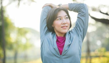 woman wearing a light blue zipper exercise top stretching her upper arm behind her facing the camera but looking away. she's in a park setting with a background of trees blurred behind her.