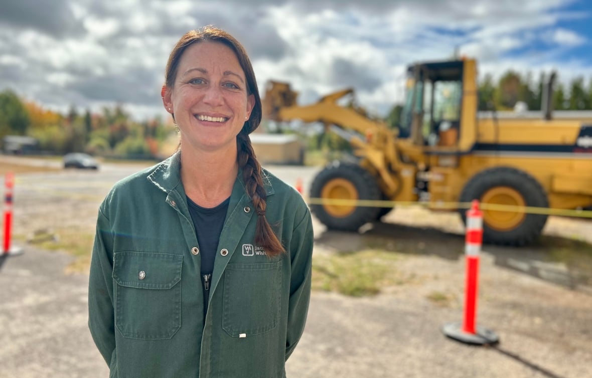 A woman with a long brown braid in her hair smiles at the camera while wearing a green jumpsuit and standing in front of a tractor. 