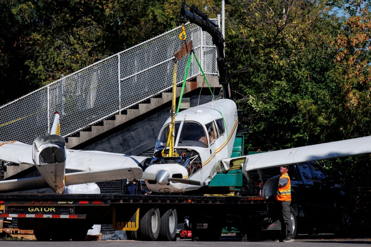 A person in reflective vest stands near a small airplane wreckage being loaded on the bed of a pickup truck, next to empty outdoor field's bleacher.