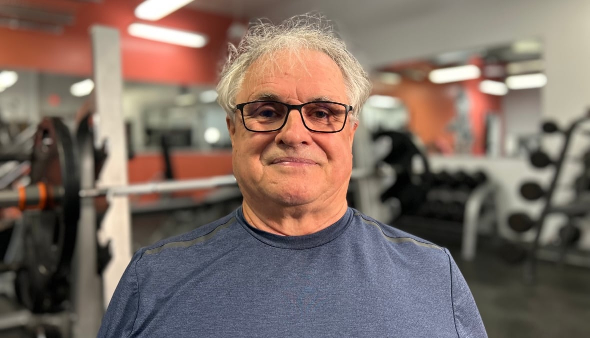 A white man with white hair, wears glasses, standing in a gym.