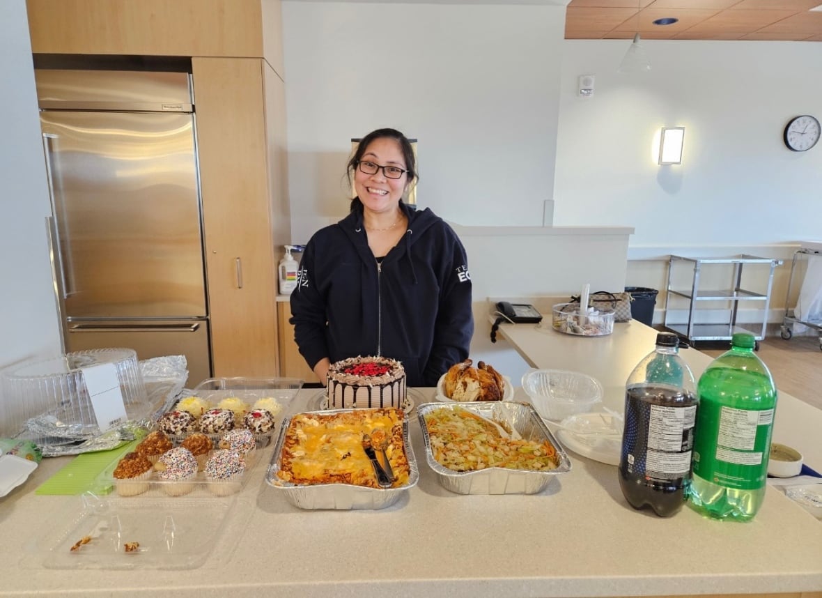 Nurse wearing glasses stands behind table that has cupcakes, Filipino food, cake, and soda bottles. 