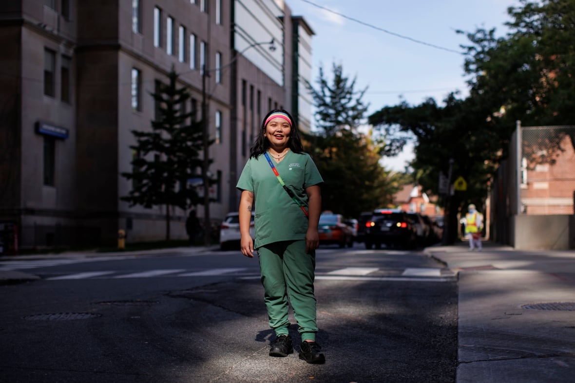 Nurse in teal scrubs and a colourful headband and bag strap standing in the middle of a street. 