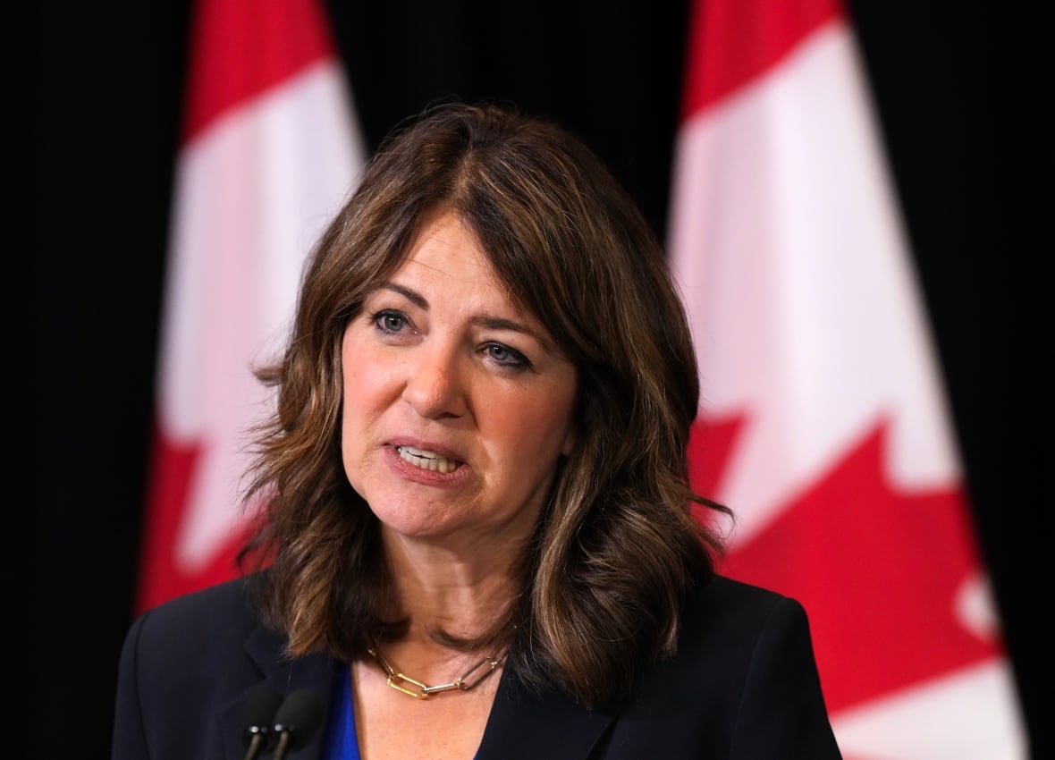Woman nearly clenches teeth while talking. Canadian flags behind her