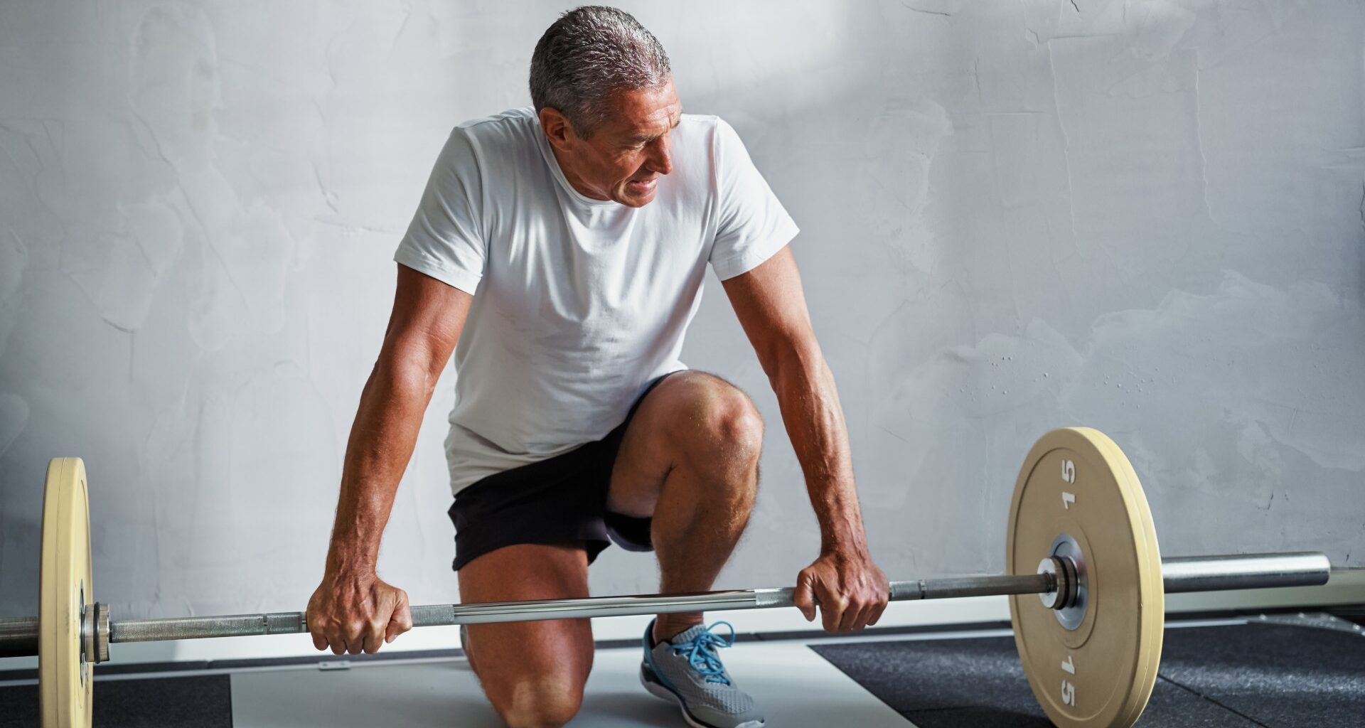 Older man in activewear crouching next to a barbell in gym