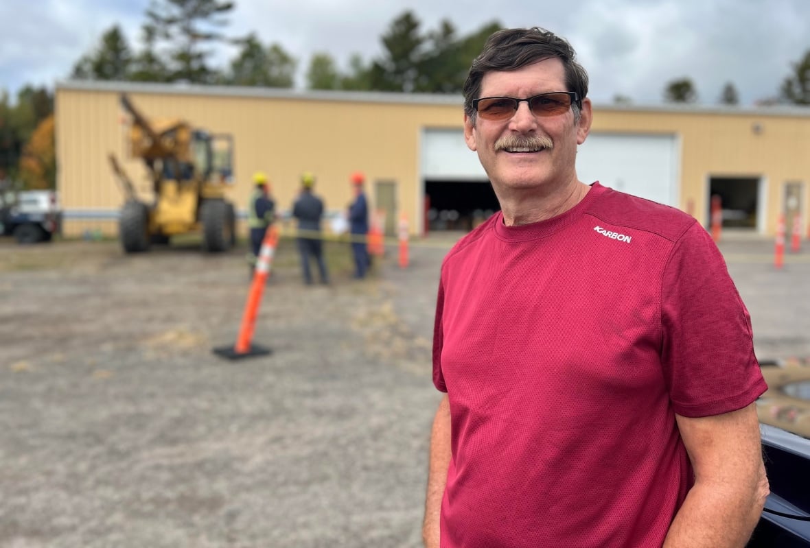 A man with brown hair and a red t-shirt smiles at the camera in front of an industrial garage with people standing out front. 