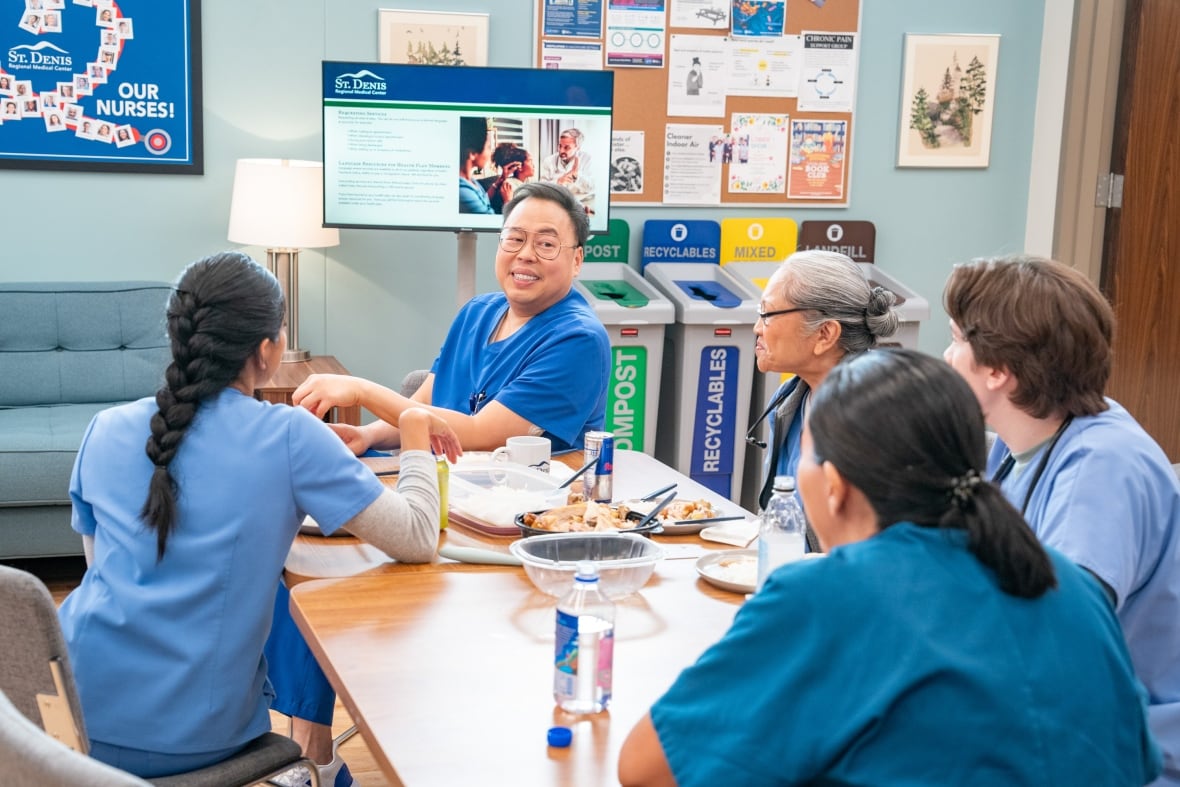 Five nurses in scrubs sit at a table in the hospital break room sharing lunch.