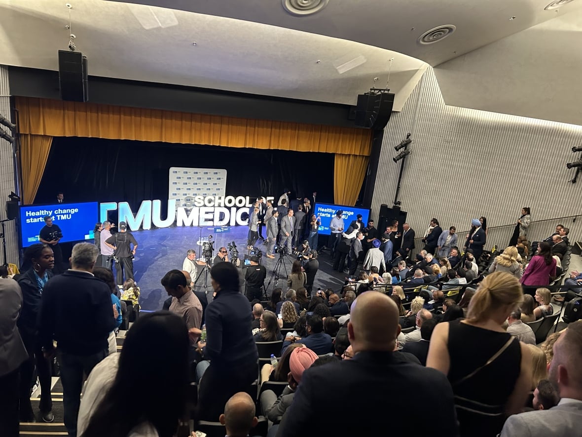 Media, reporters and people in suits attend a lecture hall for a media event.