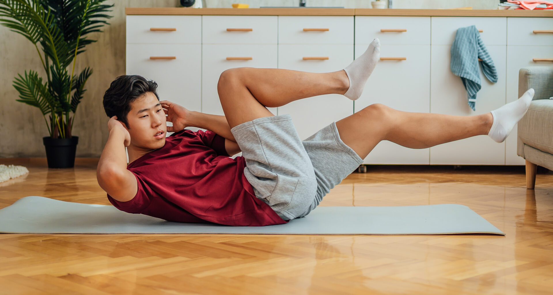 A man performs a bicycle crunch on an exercise mat at home. He is on the floor, with his lower back and buttocks in contact with the mat, while his neck, head and legs are elevated. His hands are behind his head and he is twisting to the side so that his right elbow stretches to meet his bent left knee. Behind him we see drawers and a plant.