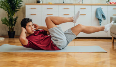 A man performs a bicycle crunch on an exercise mat at home. He is on the floor, with his lower back and buttocks in contact with the mat, while his neck, head and legs are elevated. His hands are behind his head and he is twisting to the side so that his right elbow stretches to meet his bent left knee. Behind him we see drawers and a plant.