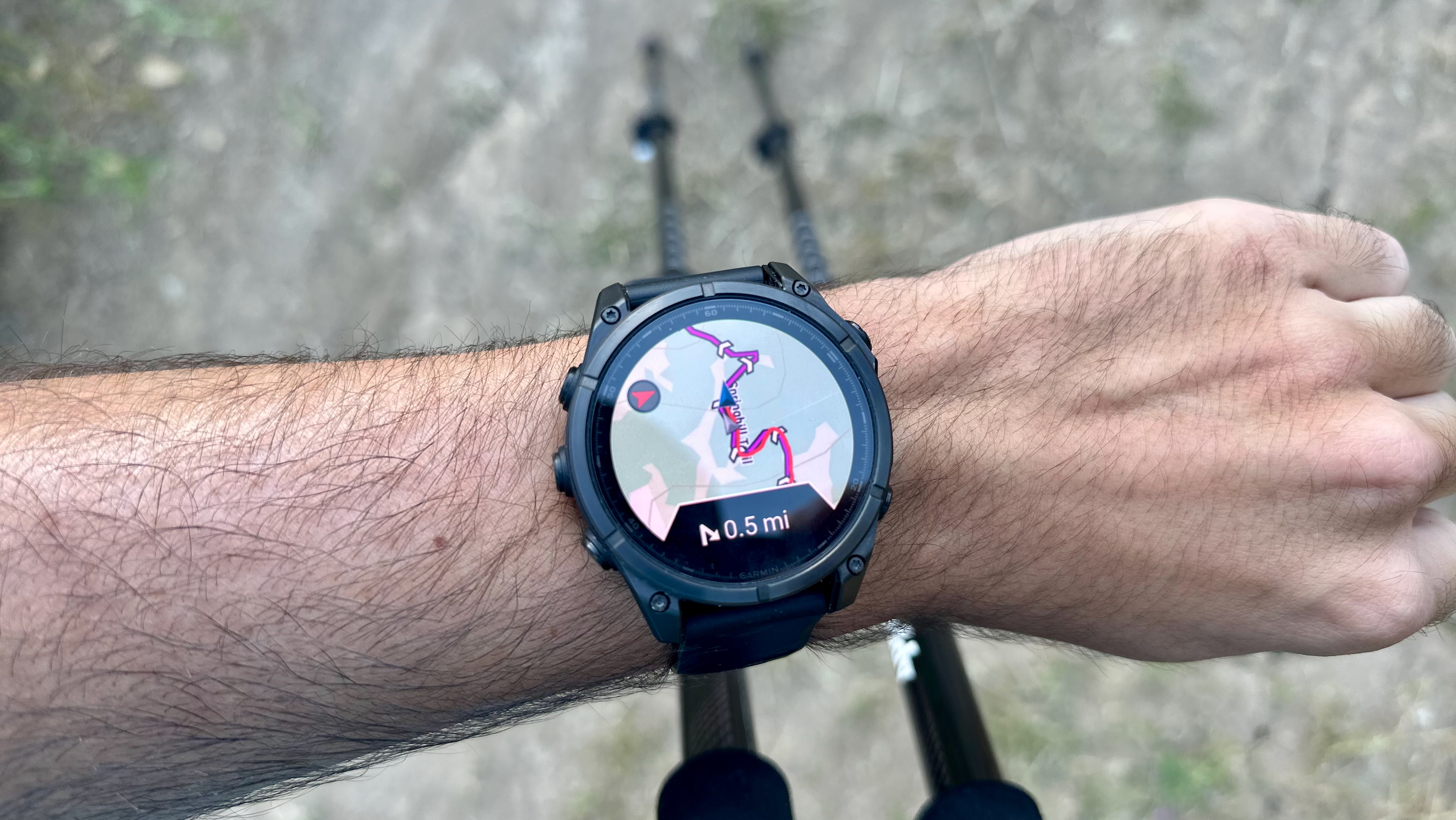 A Garmin Fenix 8 showing a course route with 0.5 miles remaining to the next turn, the watch held above a pair of trekking poles.