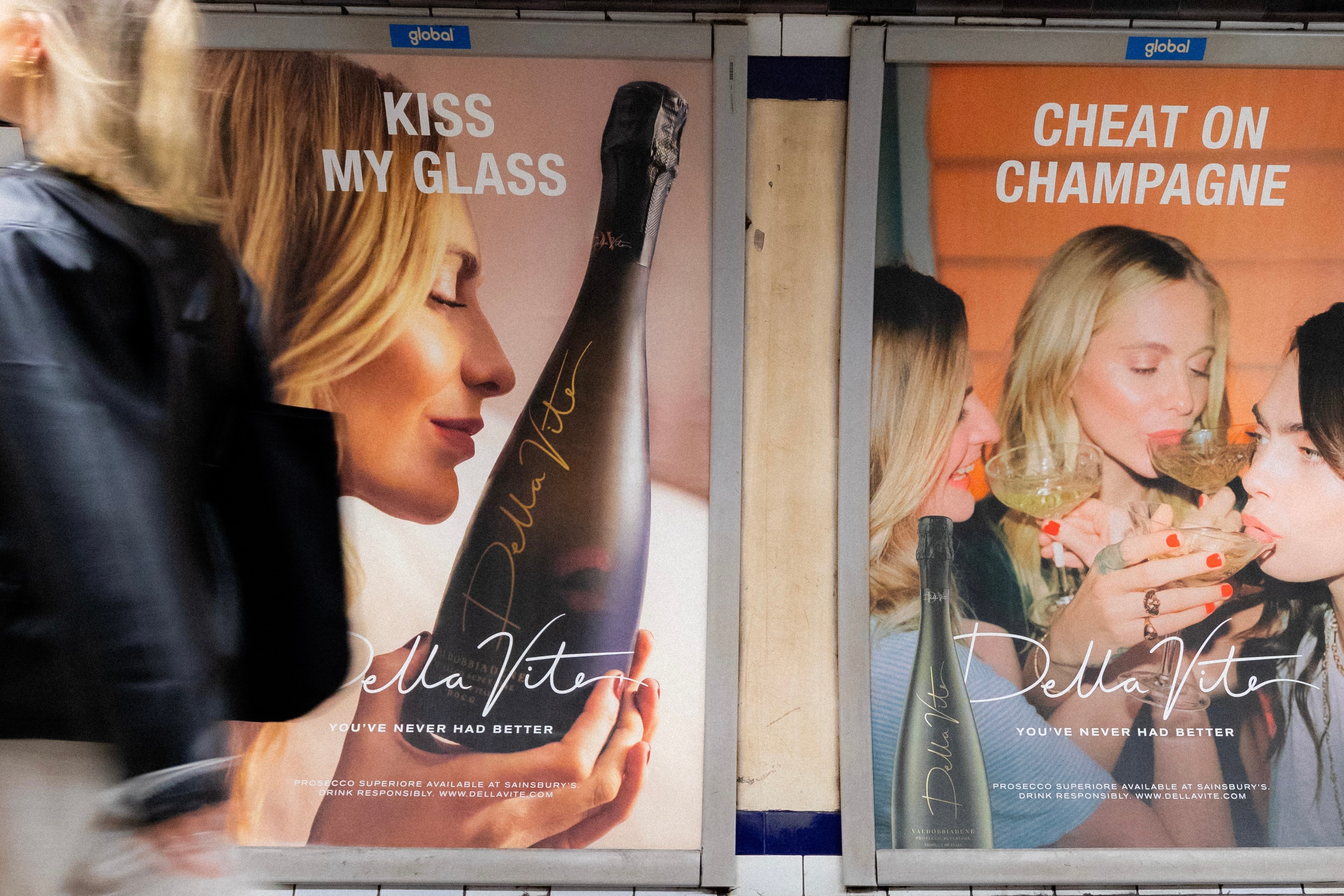Two "Della Vite" Prosecco advertisements on a subway wall, one saying "Kiss My Glass" and the other "Cheat on Champagne", with a blurred person walking past.
