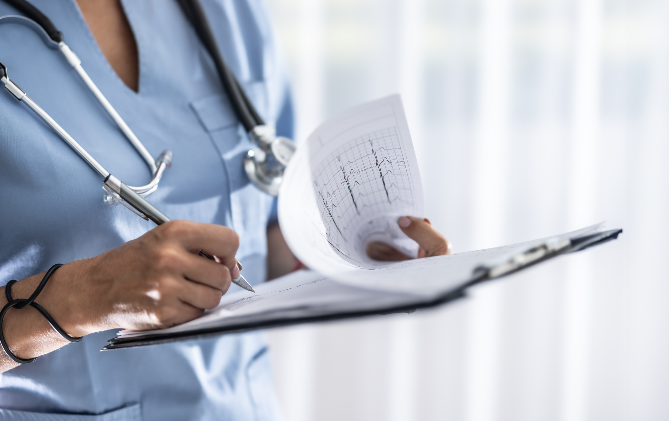 A doctor in blue scrubs signs a medical report while reviewing an EKG.