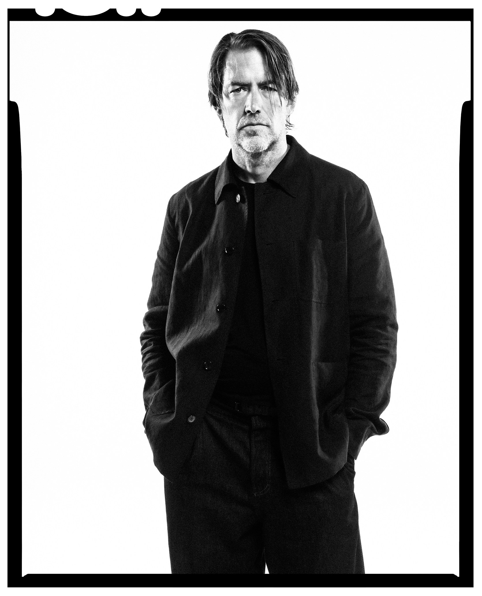 A black and white full-body portrait of a man, wearing a black shirt and black pants, with his hands in his pockets, against a white background.