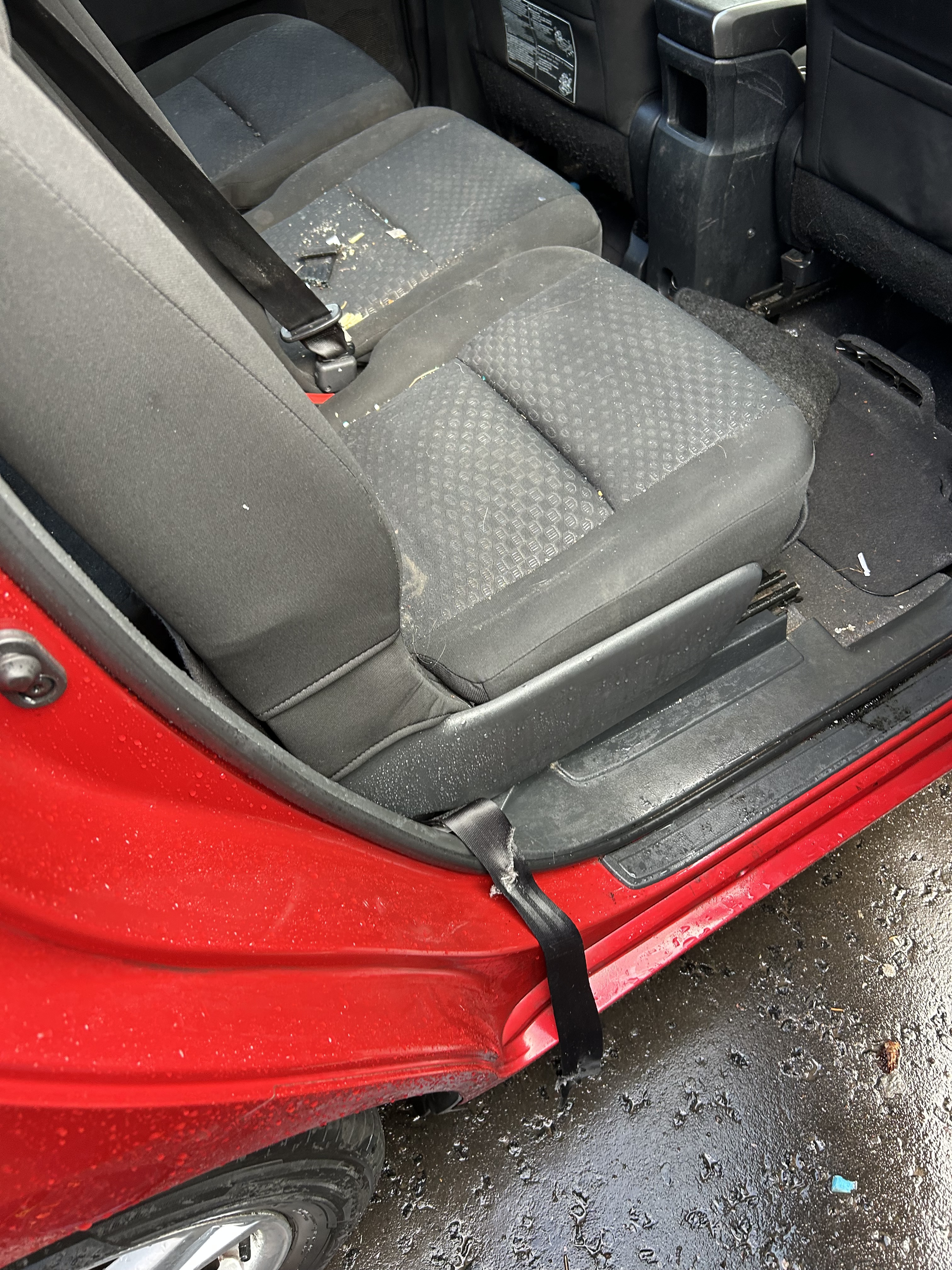 A close-up of a damaged red car's interior, showing ripped fabric on the back seat, debris on the seat and floor, and a seatbelt hanging out of the open door, which also shows exterior damage.
