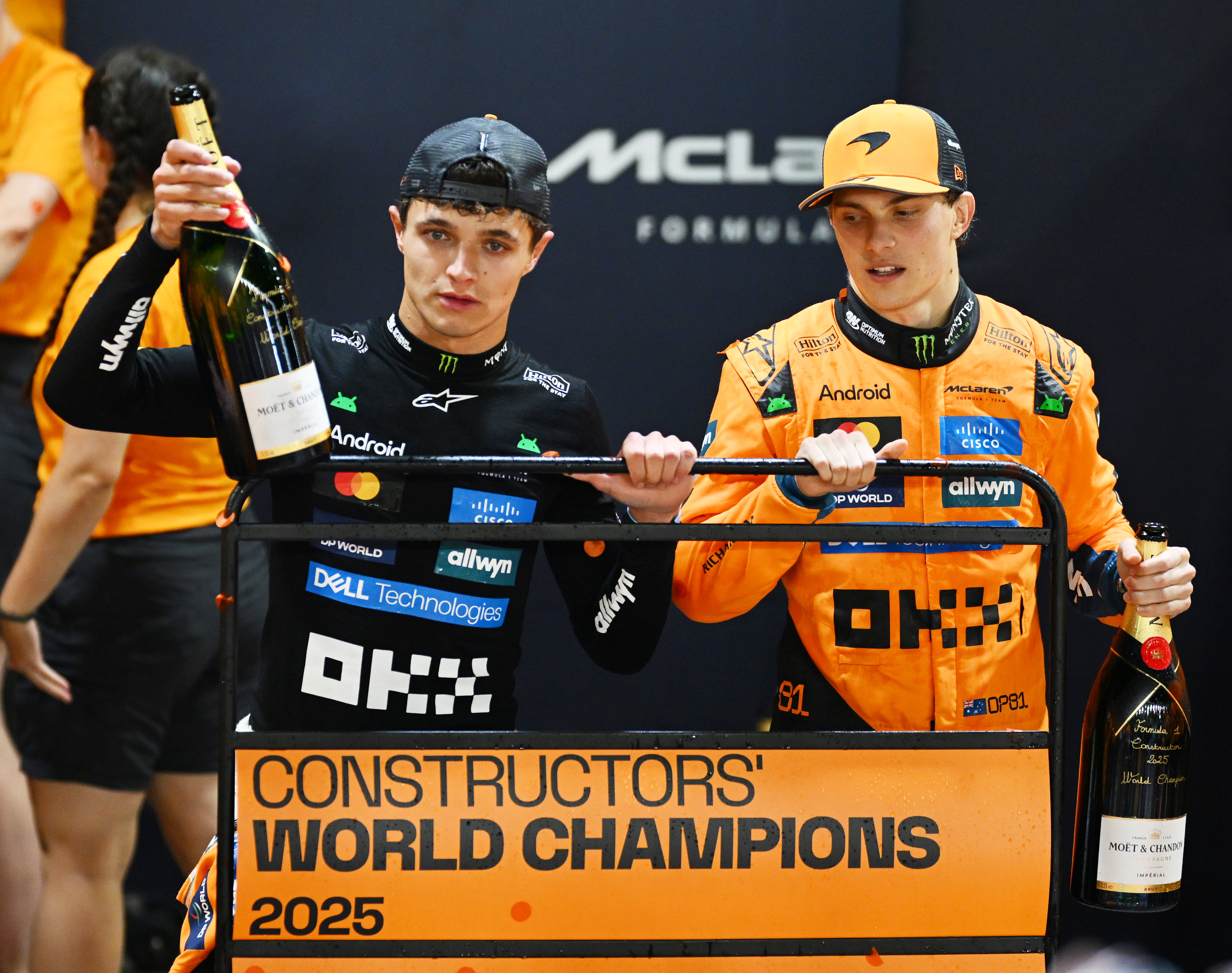 Two Formula 1 drivers, Lando Norris and Oscar Piastri, stand on a podium holding champagne bottles, celebrating as "Constructors' World Champions 2025."