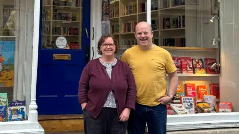 BBC Gideon and Cath York standing beside each other outside their shop,  Cotswold Book Room. Cath is wearing black jeans, a black and white spotted blouse beneath a dark purple cardigan, and has short brown curly hair and glasses. Gideon is bald and is wearing a yellow t-shirt with dark blue jeans. The shop has a dark blue front door and in the windows there are lots of books on display, lit up with warm spotlights.