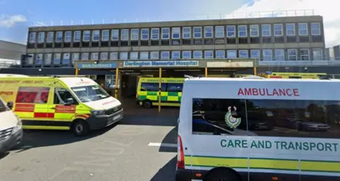 Google Several green, white and yellow ambulances parked outside Darlington Memorial Hospital. It it s a large flat grey building with two longs rows of windows above a canopy over a front door.