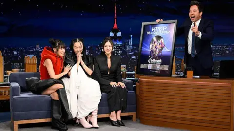 Todd Owyoung/NBC via Getty Images Rei Ami, Audrey Nuna, and EJAY, who provide the singing voices of Huntr/x. They performed on Jimmy Fallon's US talk show. Fallon is holiding a platinum disc with which the singers are being presented.