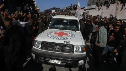 Getty Images A red cross vehicle driving through a crowd of people. It is white with the red cross on the front. 