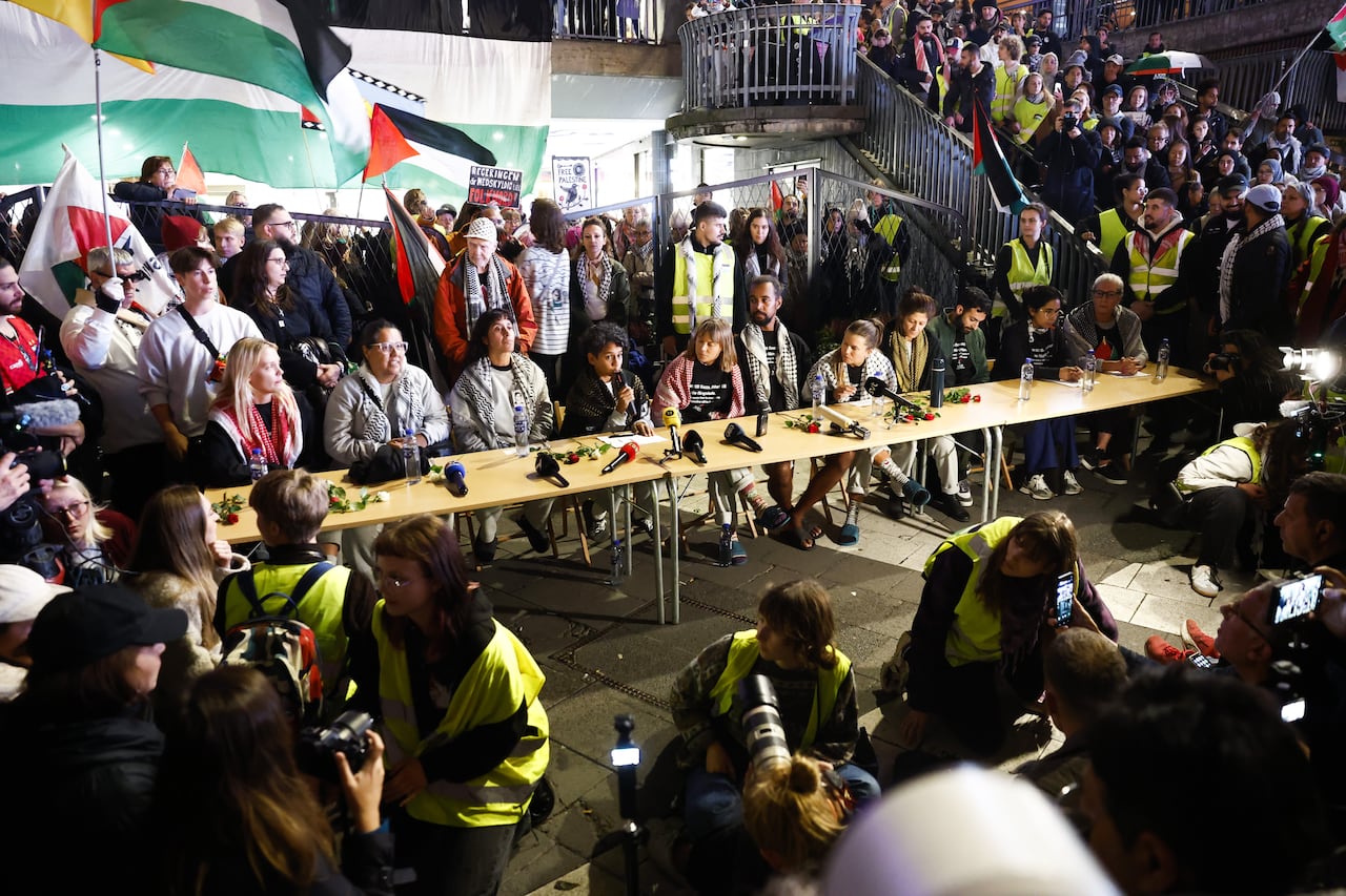 A group of people sit along a panel as people gather around them.