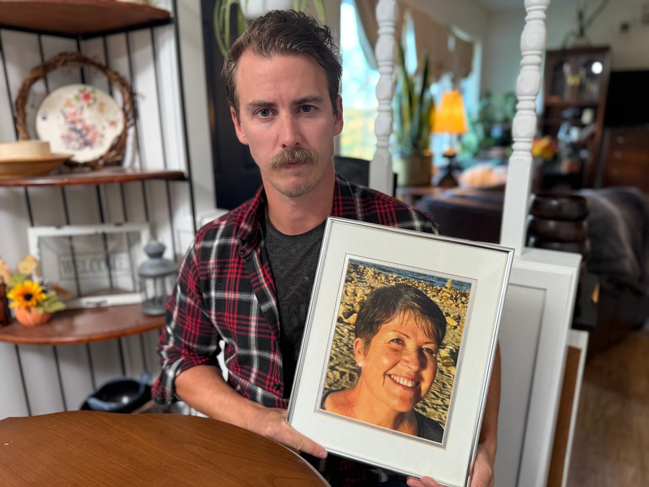 A man in his 30s with a moustache sits at his kitchen table, looking into the camera, holding a framed photo of a 69-year-old woman