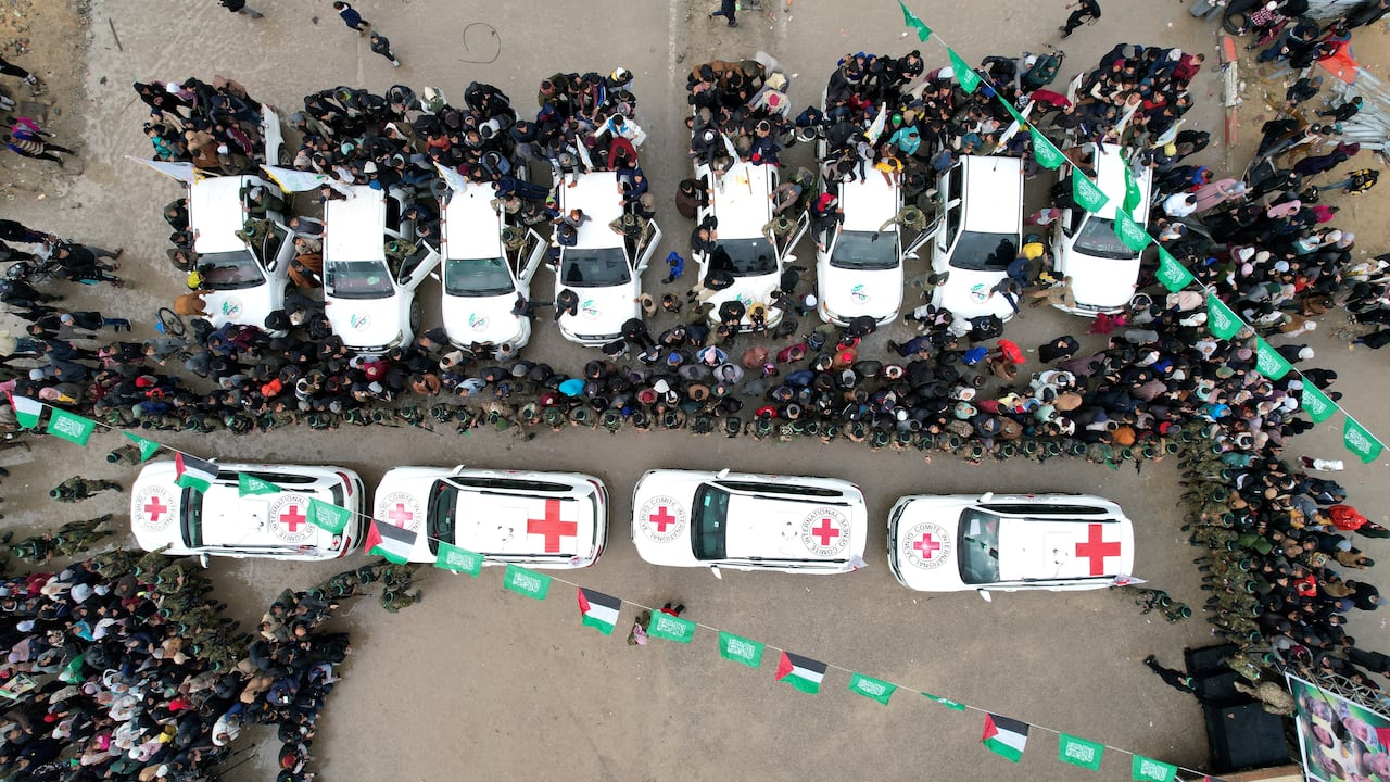 A photo taken from above shows a line of white vehicles with red crosses on them driving through a  crowd