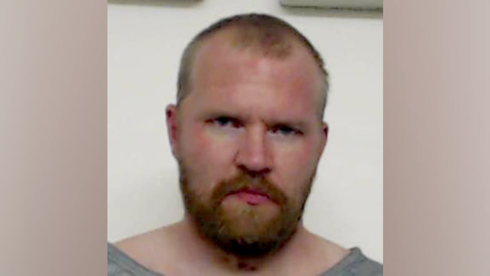 A man is seen from the shoulders up. He has a beard and a closely cropped haircut. He is not smiling.