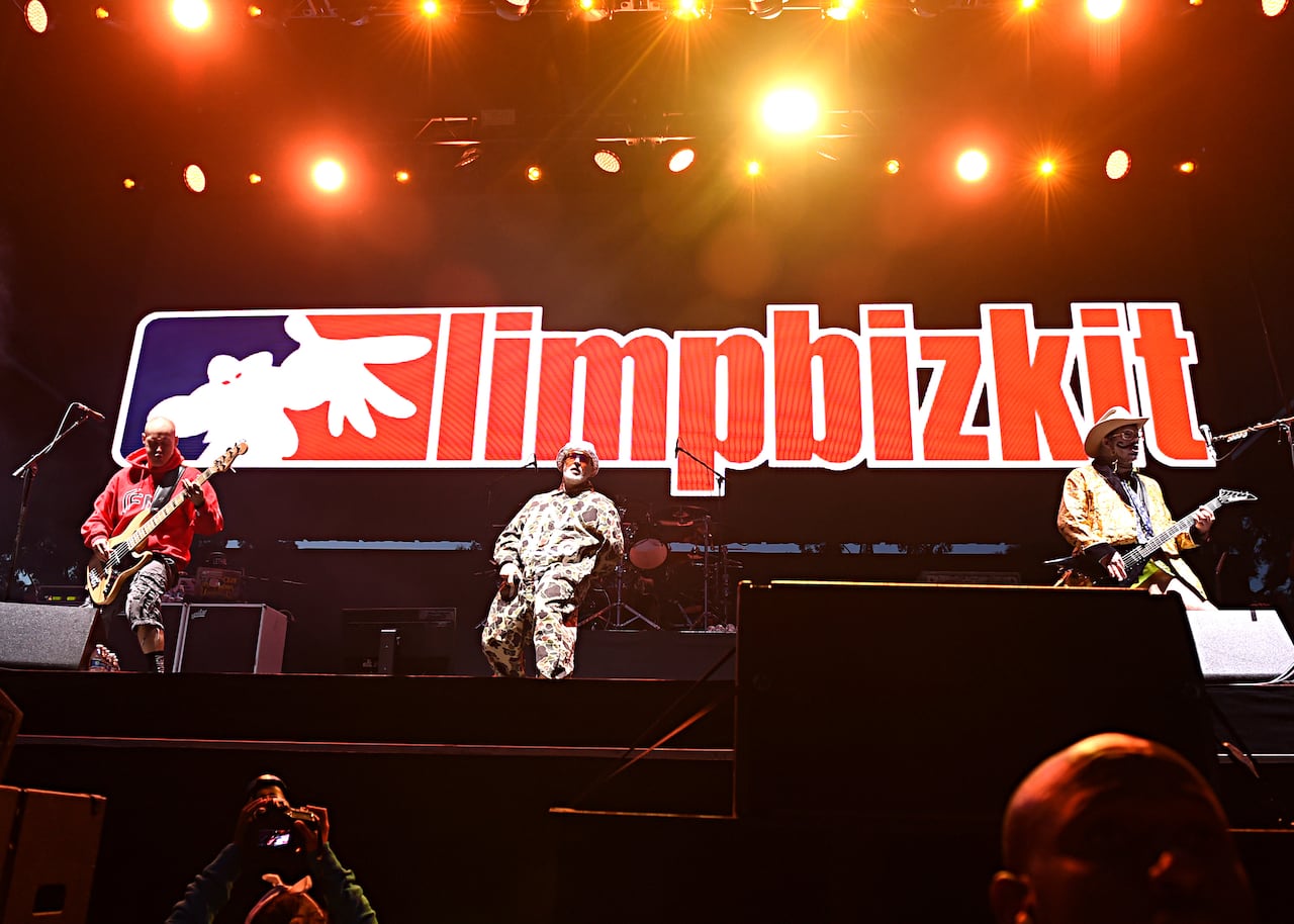Three men perform on a stage at night with a screen behind showing the band name Limp Bizkit in large red letters.