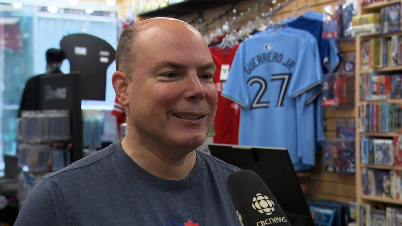 A smiling man wearing a Toronto Blue Jays shirt stands in a sports shop.