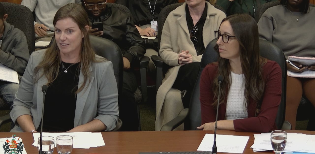 Two women sit at a table speaking into microphones, with a crowd of people sitting behind them