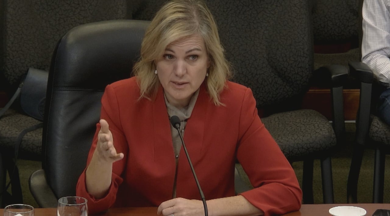 A woman wearing a red blazer speaks into a microphone mounted onto a table