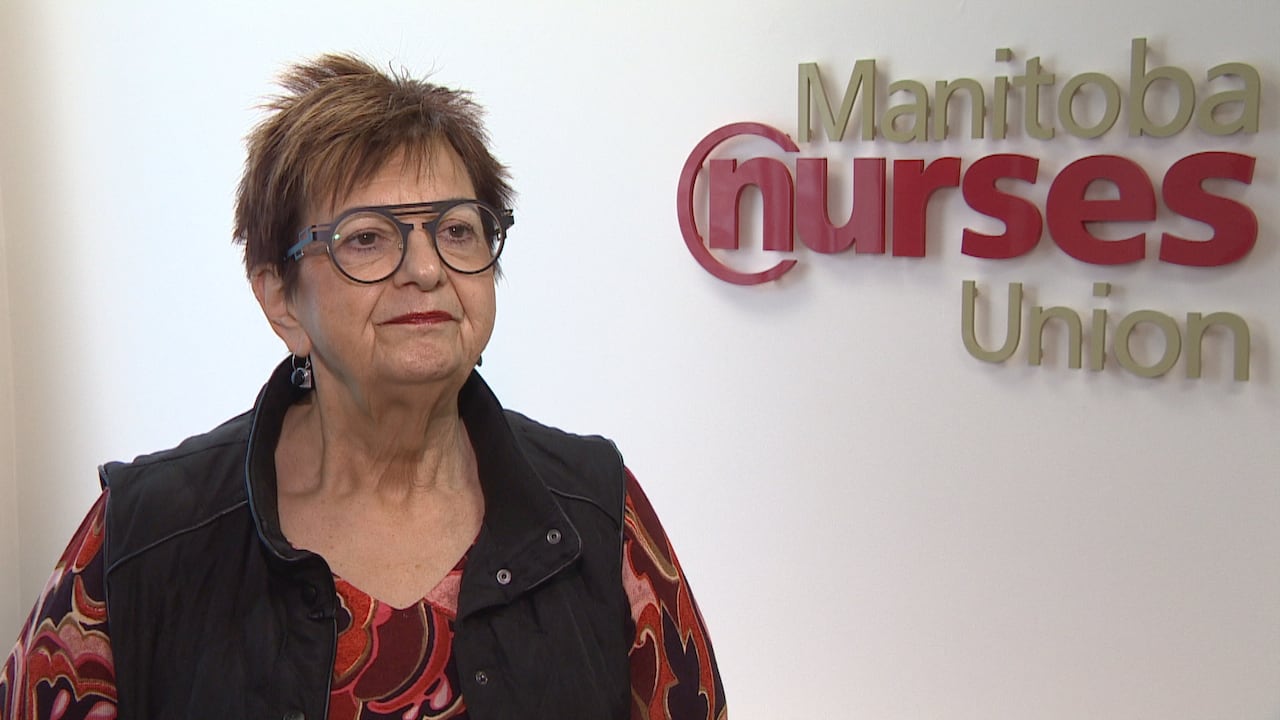 A woman with short hair and glasses stands facing a camera