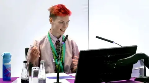 Lampard Inquiry/YouTube Hat Porter gives evidence to the Lampard Inquiry. They are wearing a light coloured button-down shirt with a green tie, with several lanyards around their neck. They have a short haircut which is dyed bright red. They are sat at a purple table with two microphones and a computer monitor in front of them