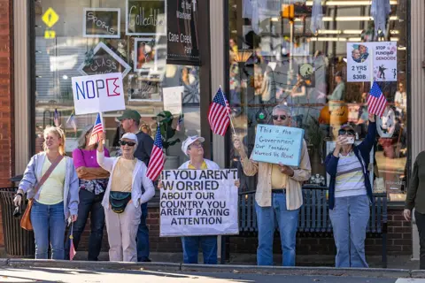 Getty Images A small group of protestors is gathered outside a shop. They carry American flags and carry signs that read "If you aren't worried about our country you aren't paying attention" and "This is the government our founders worried about"