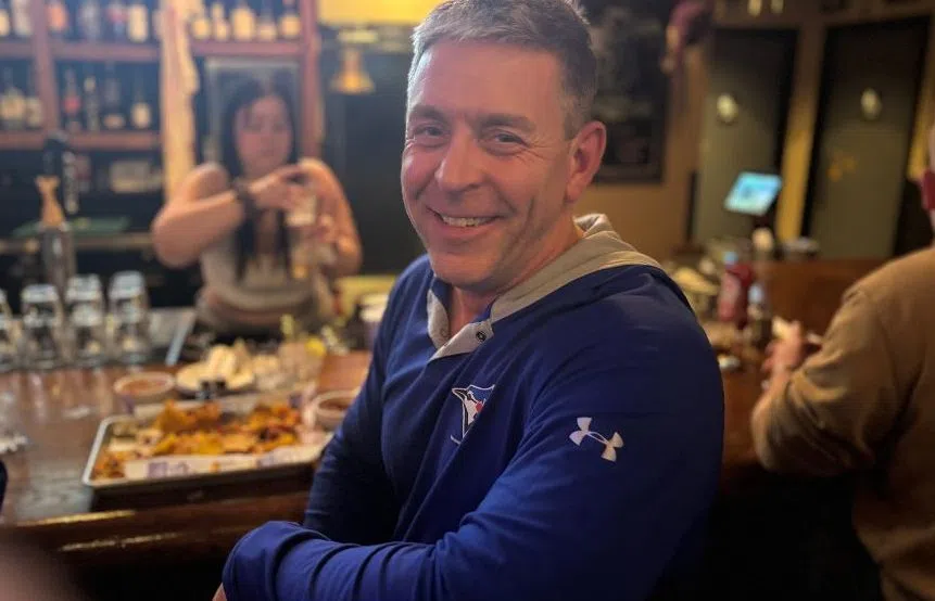 A man wearing a Blue Jays hoodie smiles at a bar table covered with food and drinks while watching the game on TV.