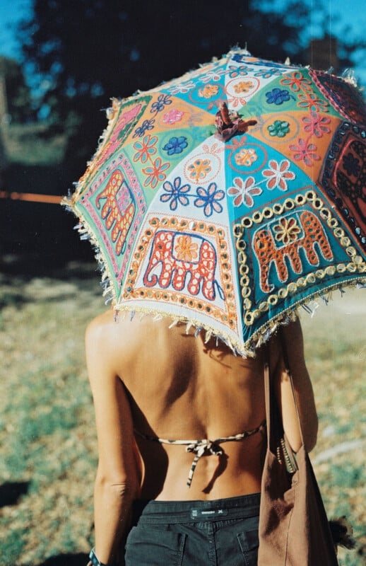 A person in a bikini top and black pants stands outdoors, holding a colorful, embroidered umbrella decorated with elephants and floral patterns, with sunlight casting shadows on their back.