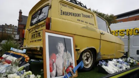 Reuters The famous yellow van from Only Fools And Horses is on the back of a truck. It is surrounded by flowers. 