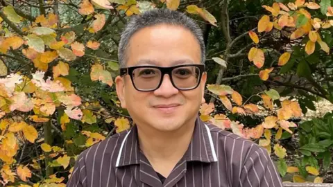 NWAFT Angelo wearing a brown and white stiped nurse uniform and glasses - smiling for the camera. There are bushes 