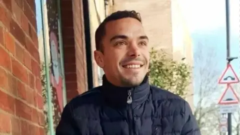 PA A smiling Leonard Farruku looks off to the side of the camera. He has smartly cropped dark hair and a stubble goatee beard. He is wearing a navy blue padded winter jacket, zipped up to his chin.