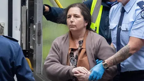 Reuters Erin Patterson being walked along by security officers, in handcuffs. She's wearing a pattered shirt with a two brown cardigans over the top. She has brown and grey hair tied back.