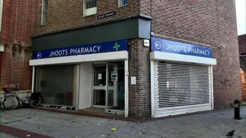BBC A closed down pharmacy. The name reads JHOOTS pharmacy in white writing on a blue background above the shop. The shutters are down covering the window to the right.