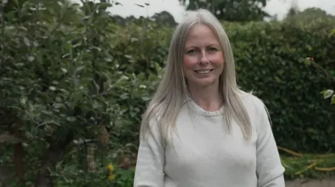 Andy Alcroft/BBC Emily, wearing a cream jumper, smiles as she stands in front of hedges and a tree with a bird feeder hanging in its branches